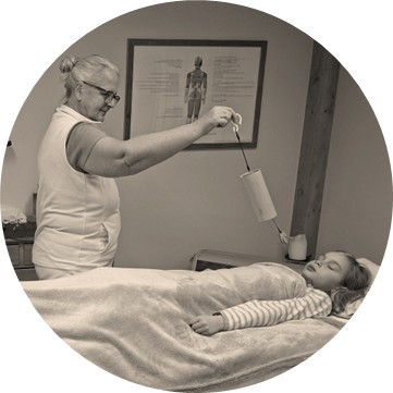 Phonophorese (tuning fork therapy) close-up: therapist’s hands holding vibrating tuning forks above the forearm, soft warm light, shallow depth of field, calming wellness studio, realistic photo